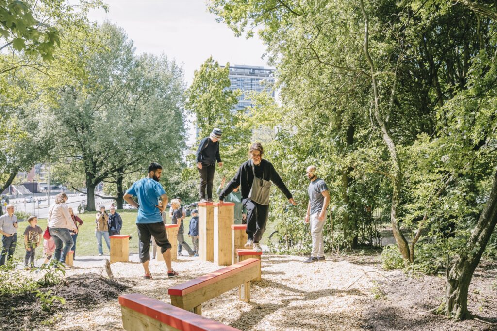 Menschen unterschiedlichen Alters nutzen in einem Park eine Holz-Kletter- und Balancierstation zwischen Bäumen.