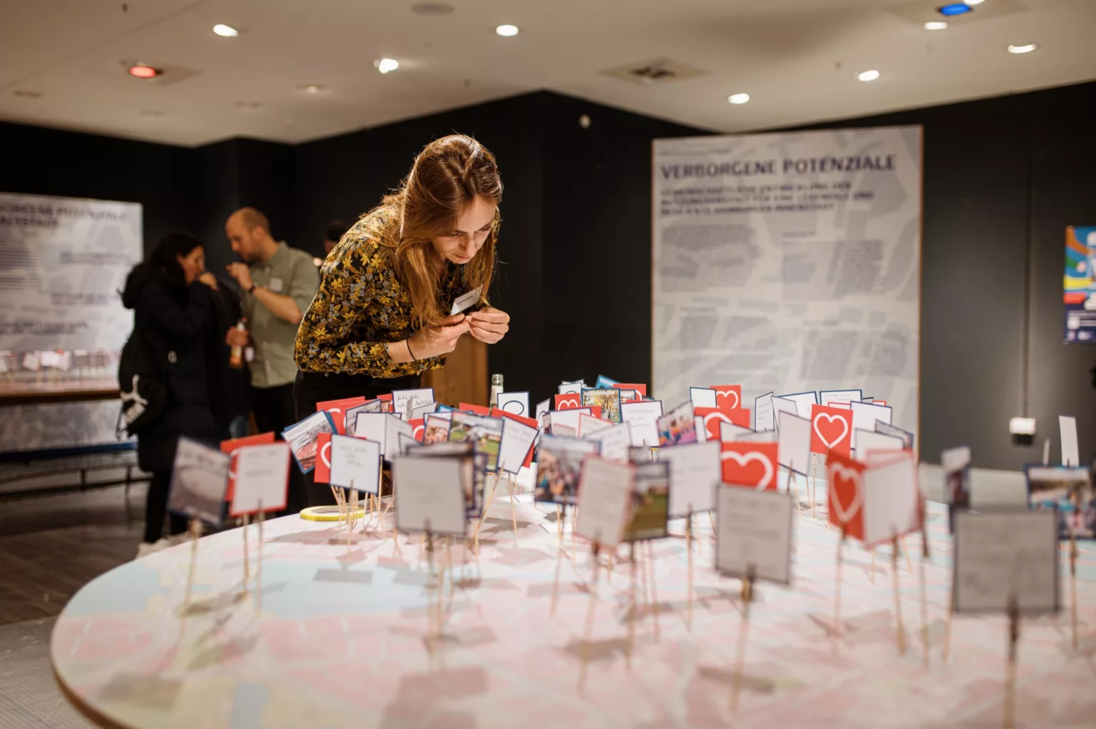Frau beugt sich über einen großen Stadtplan mit zahlreichen kleinen Schildchen und Symbolkarten, die auf Holzstäbchen befestigt sind, im Rahmen einer Stadtwerkstatt-Ausstellung.
