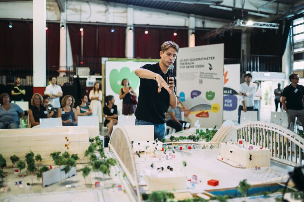 Junger Mann spricht mit Mikrofon vor einem Publikum und diskutiert ein das Modell zu dem Projekt, in der Halle um die es geht.