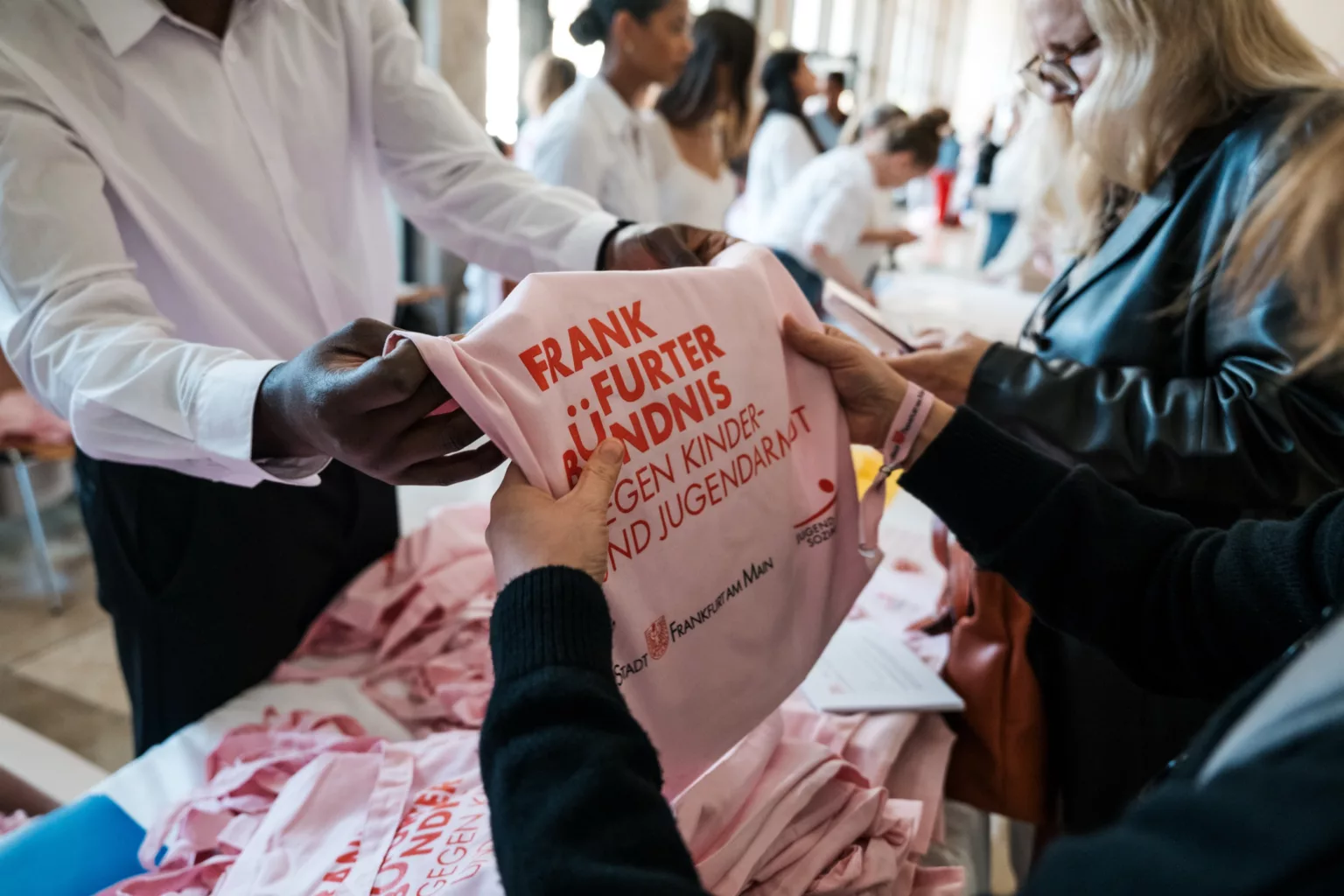Nahaufnahme einer Person, die einer anderen eine rosa Stofftasche mit der Aufschrift ‚Frankfurter Bündnis gegen Kinder- und Jugendarmut‘ überreicht. Im Hintergrund stehen weitere Menschen an einem Tisch mit mehreren gleichartigen Taschen.