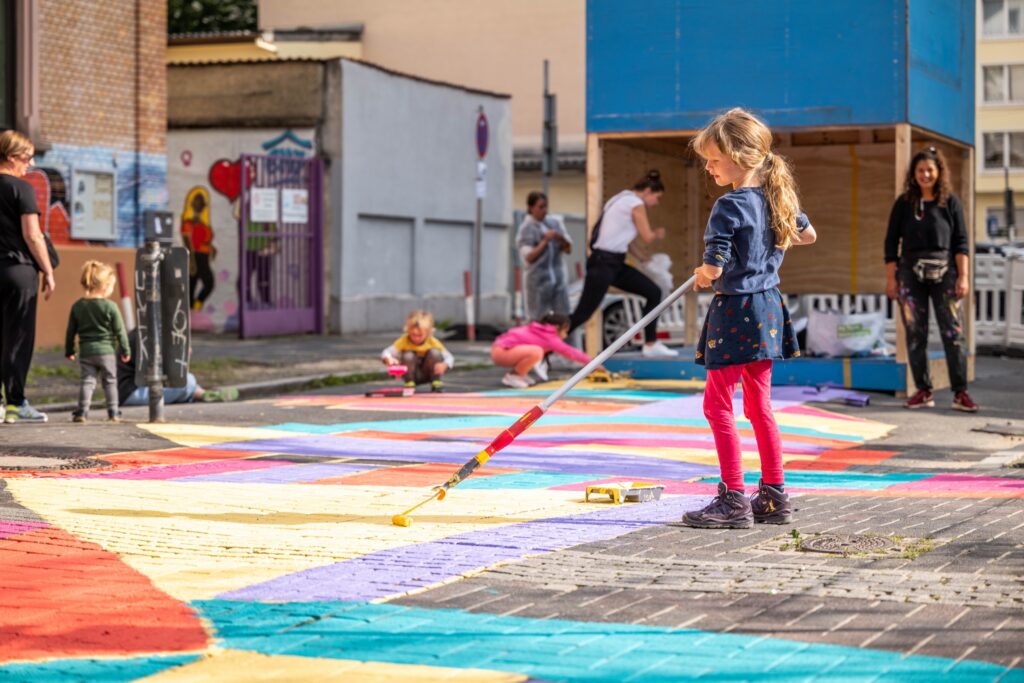 Kinder bemalen gemeinsam mit Erwachsenen eine Straße bunt mit Farbe im Rahmen einer Mitmachaktion im öffentlichen Raum.