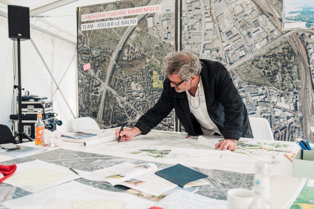 Ein Mann zeichnet einen Plan auf Transparenzpapier. Hinter ihm ist ein großes Luftbild von Bahrenfeld, Hamburg.