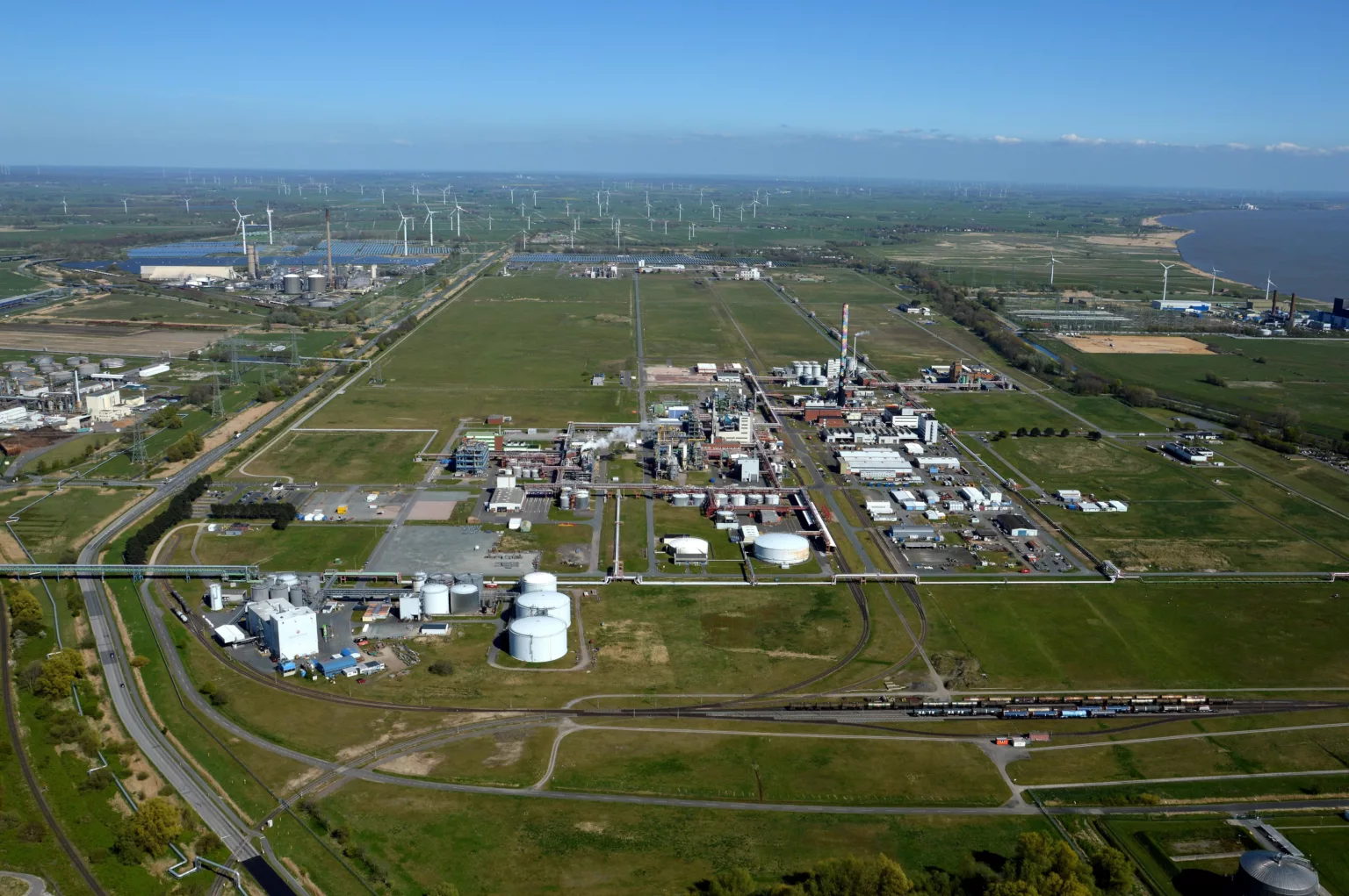 Luftaufnahme des Industrie- und Energieparks in Brunsbüttel mit Industrieanlagen umgeben von Rasenflächen. Im Hintergrund eine Vielzahl an Windrädern und die Elbe.