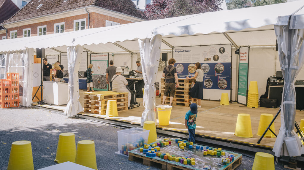 Ausstellungszelt mit Infotafeln und Palettentischen, davor spielt ein Kind mit bunten Bauklötzen auf einem Stadtplan.