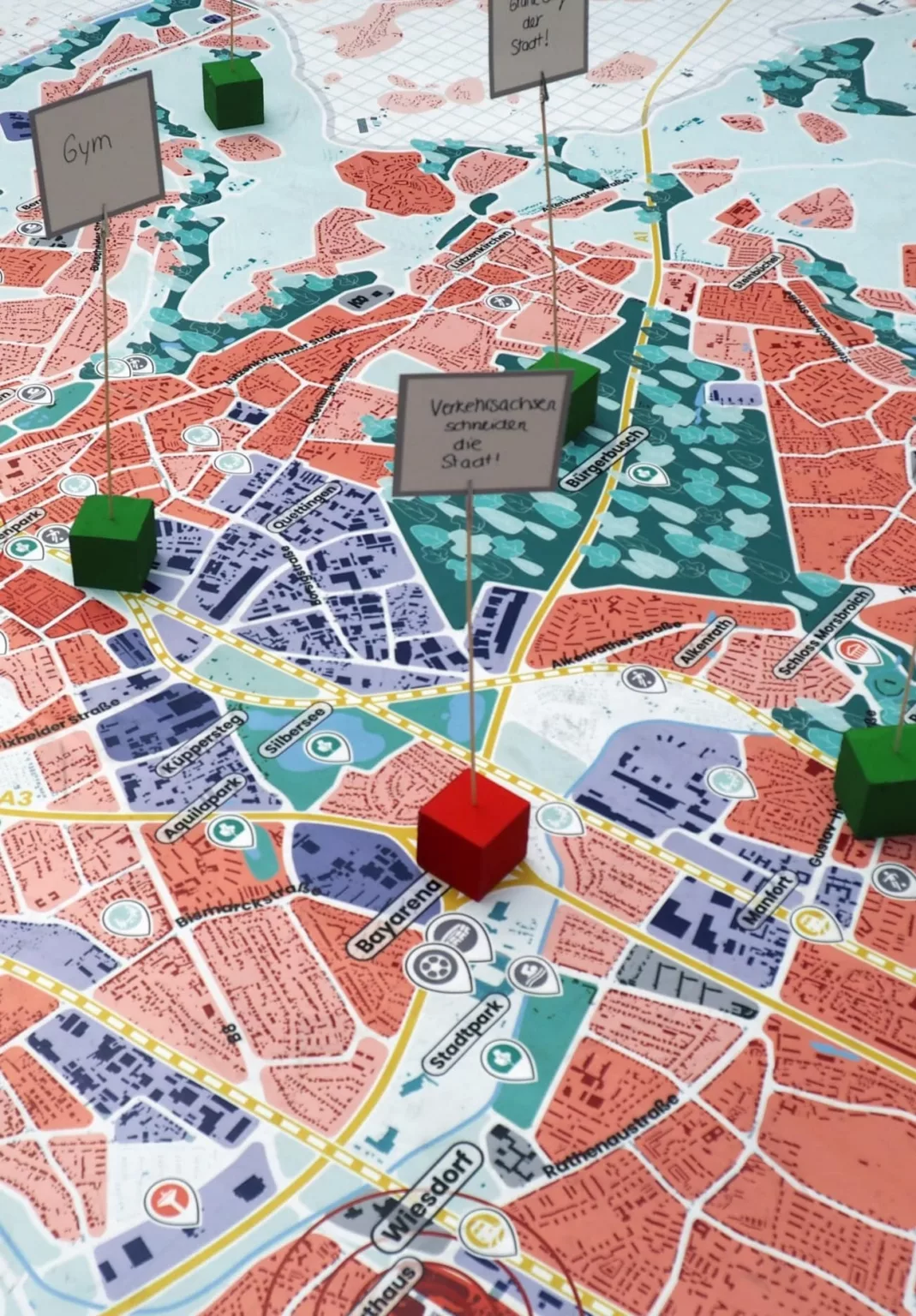 Stadtkarte mit farbig markierten Gebieten, Straßennetzen und Flüssen. Darauf stehen mehrere rote und grüne Würfel mit kleinen Schildern, auf denen handschriftliche Notizen zu Orten und Themen wie ‚Gym‘ angebracht sind.
