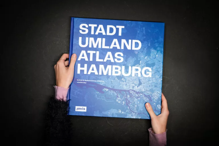 Person hält ein Buch mit dem Titel Stadt Umland Atlas Hamburg vor schwarzem Hintergrund.