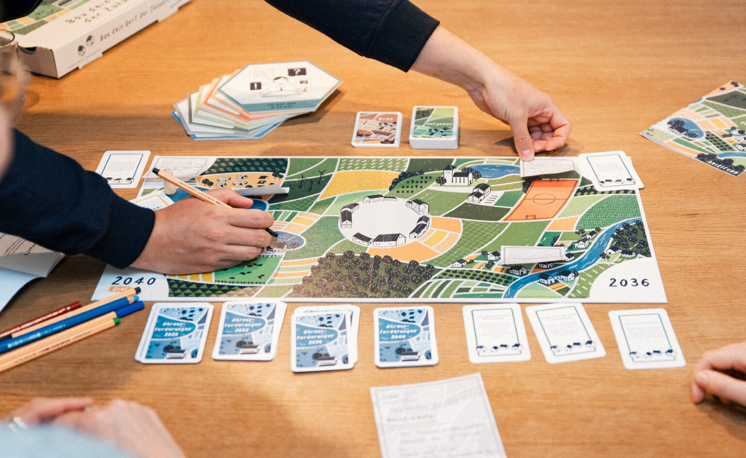 Es wird das von urbanista entwickelte Spiel Bau dein Dorf der Zukunft gespielt. Zu sehen sind der Spielplan und Spielkarten.