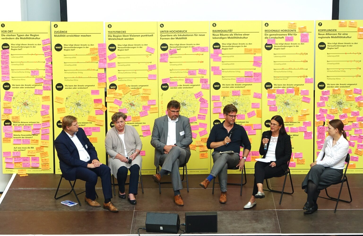 Eine Podiumsdiskussion zur IBA München mit fünf Teilnehmenden. Im Hintergrund zu sehen sind Workshop-Ergebnistafeln zu sieben Themen